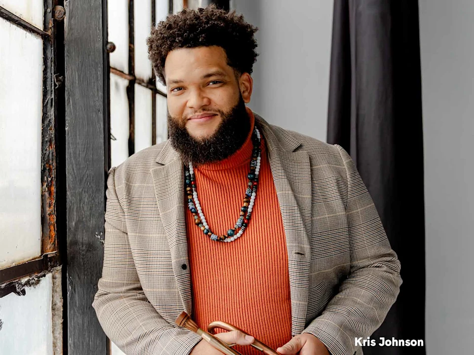 A man with a beard and curly hair, wearing a plaid blazer, orange sweater, and beaded necklaces, stands by a window holding an object. The name "Kris Johnson" appears in the bottom right corner.
