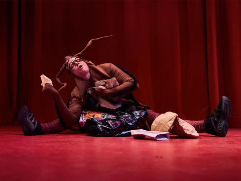 A person in a bug costume sits on stage with a red curtain backdrop, surrounded by various paper and plastic bags.