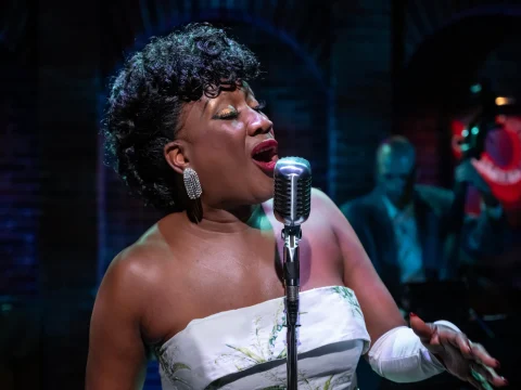 Person in a white dress sings passionately into a vintage microphone on stage, with a musician playing an upright bass in the background.