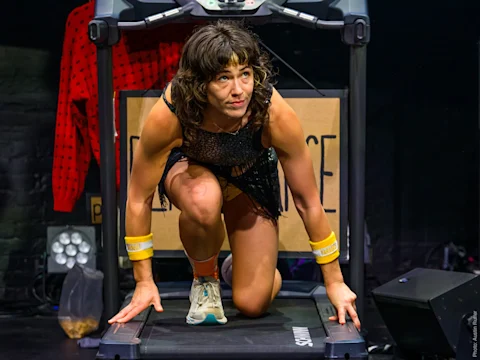 A woman kneels in a runner’s starting position on a treadmill, wearing athletic gear and wristbands, with stage lighting and props visible in the background.