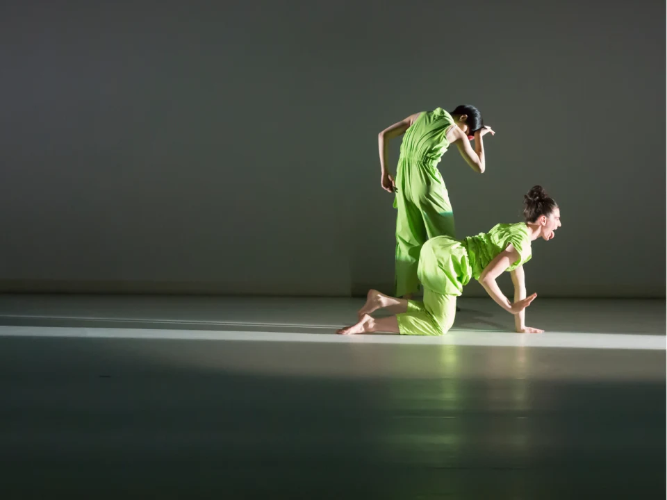 Two dancers in green outfits perform on a stage, one standing and the other on hands and knees, both illuminated by a spotlight.