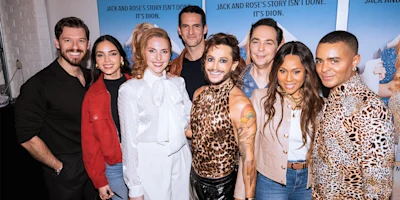 A group of eight people stand together smiling for a photo in front of posters with text in a brightly lit indoor setting.