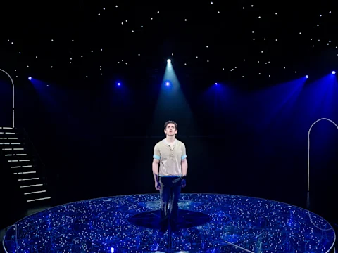 A man stands alone on a circular, illuminated stage with blue lighting and a star-like pattern, under a spotlight, with stairs and archways in the background.