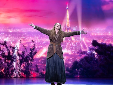 A performer stands center stage with arms outstretched, singing, in front of a vibrant Paris skyline backdrop featuring the Eiffel Tower at dusk.