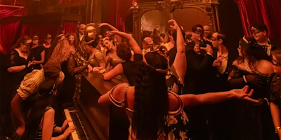 A group of people in formal attire and masks gather around a piano in a dimly lit, ornate room with red curtains, some dancing and posing in theatrical fashion.