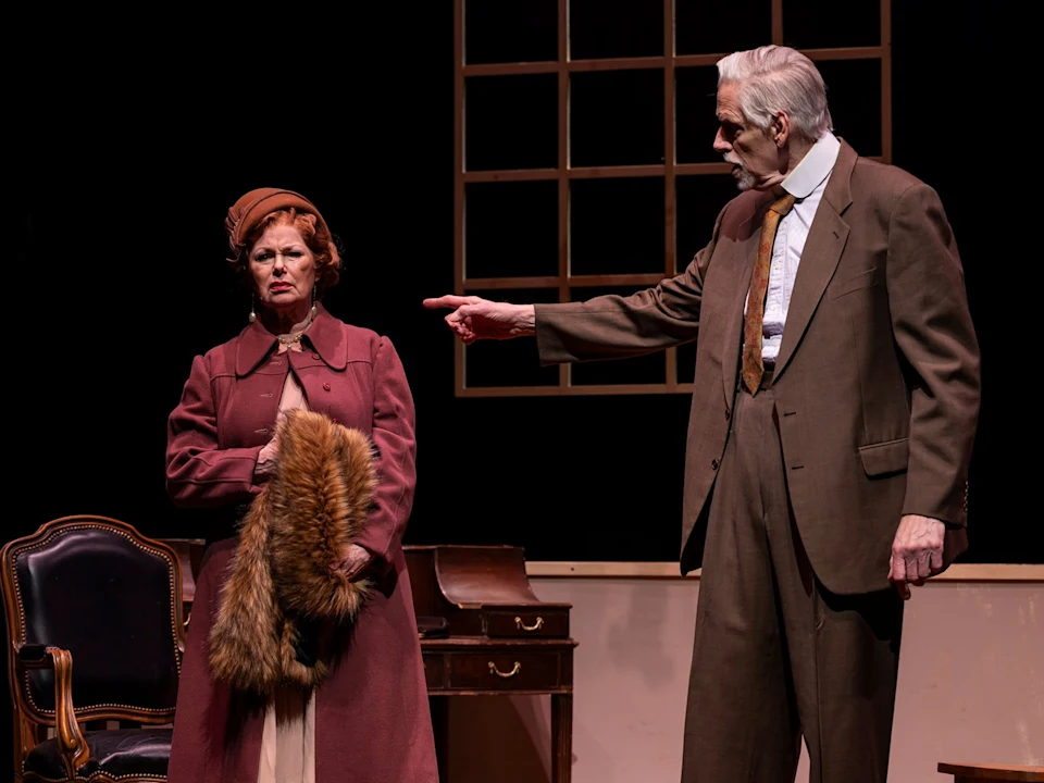 An older man in a suit points sternly at a woman in a maroon coat and hat, who stands with a fur stole; they are on a stage set with office furniture.