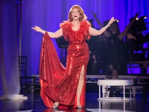 A performer in a red sequin dress with a feather boa sings on stage with a spotlight, while musicians play in the background.