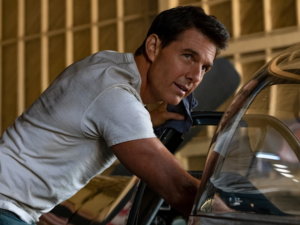 A man in a white t-shirt leans on a cockpit, holding a cloth, with a hangar background and natural light.