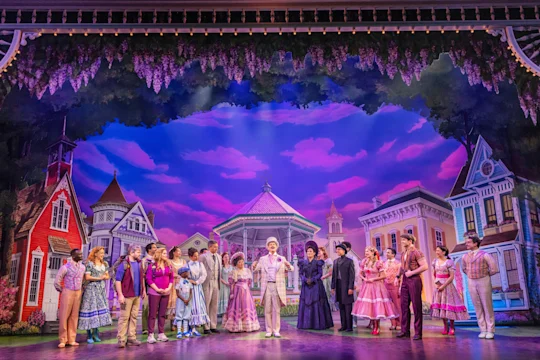 A large cast in colorful period costumes stands on stage with painted Victorian house backdrops and a sky-blue background, performing a theatrical musical scene.