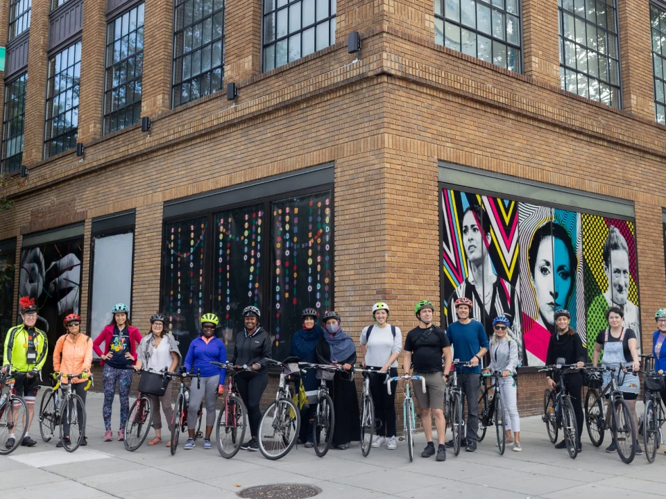 A group of people with bicycles stand in front of a brick building with large colorful murals on the windows.