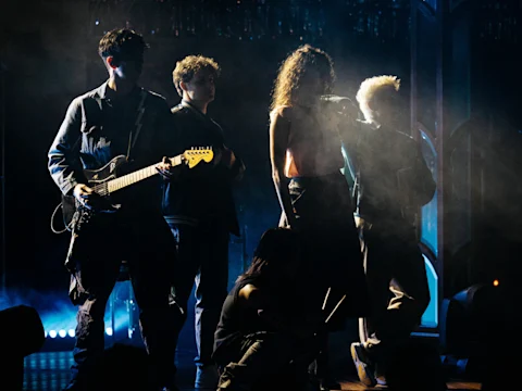 Four musicians stand on a dimly lit stage, one holding an electric guitar. Smoke and blue backlighting create a dramatic atmosphere.