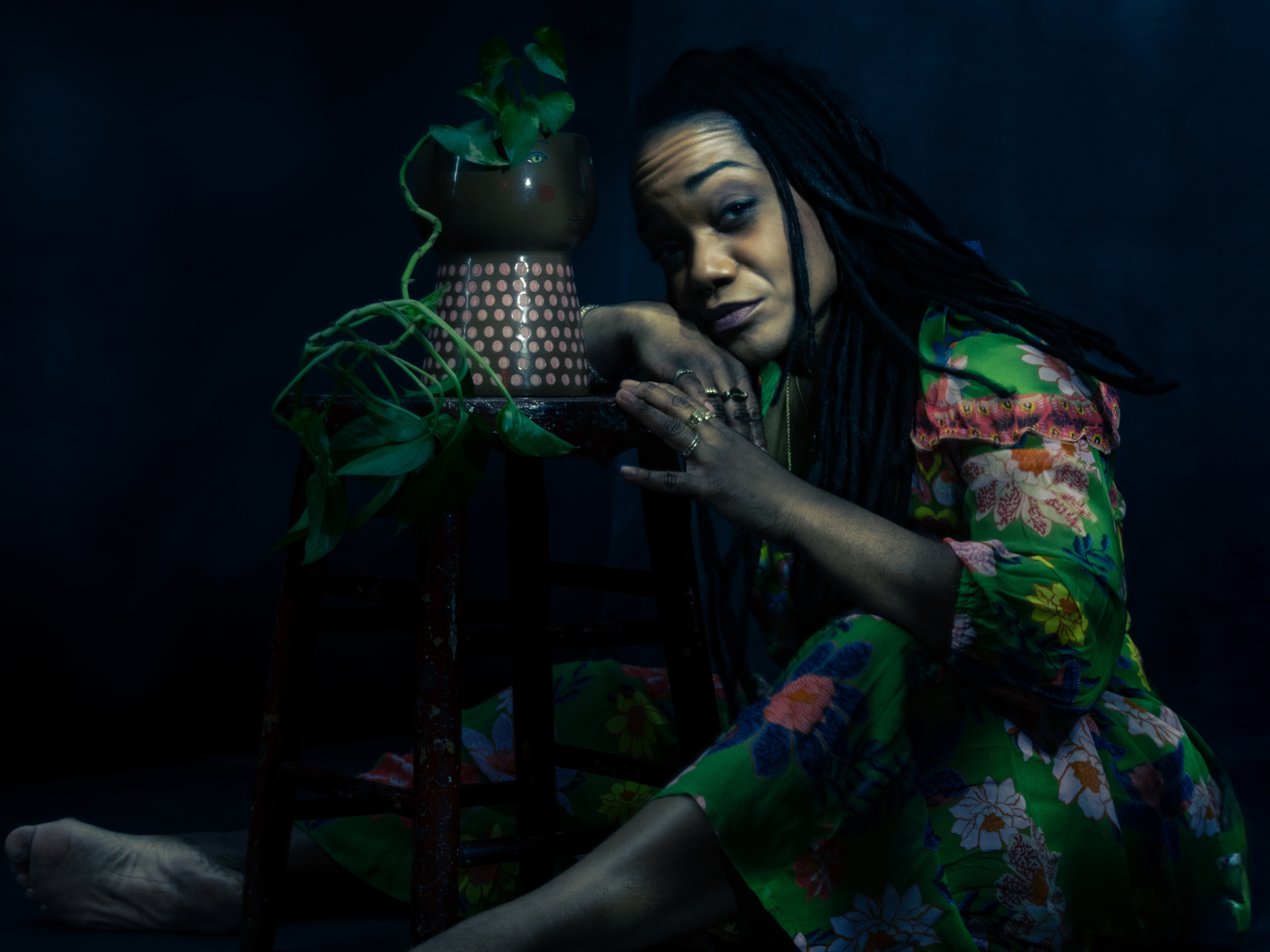 A woman in a floral dress sits barefoot on the floor next to a stool with a plant in a pot, looking directly at the camera in dim lighting.