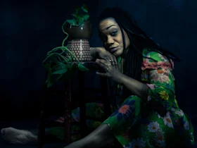 A woman in a floral dress sits barefoot on the floor next to a stool with a plant in a pot, looking directly at the camera in dim lighting.