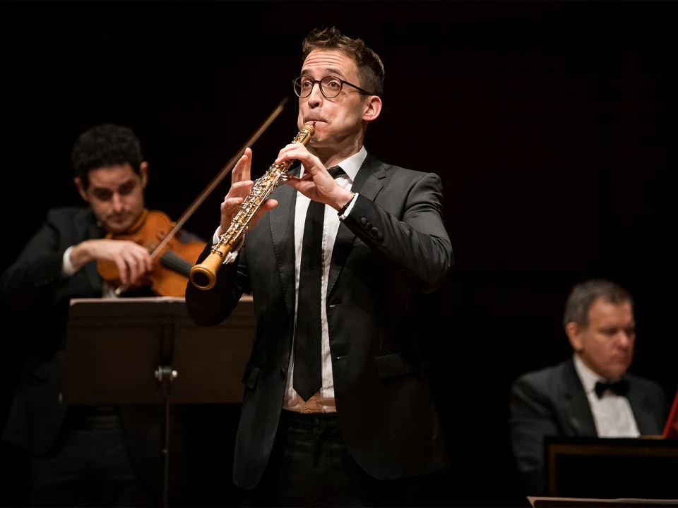 A man in a suit plays an oboe on stage, accompanied by a violinist and another musician in the background.