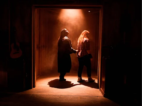 Two people stand in a dimly lit doorway with mist and a spotlight shining down, one person holding the other by the arm as they face away from the camera.