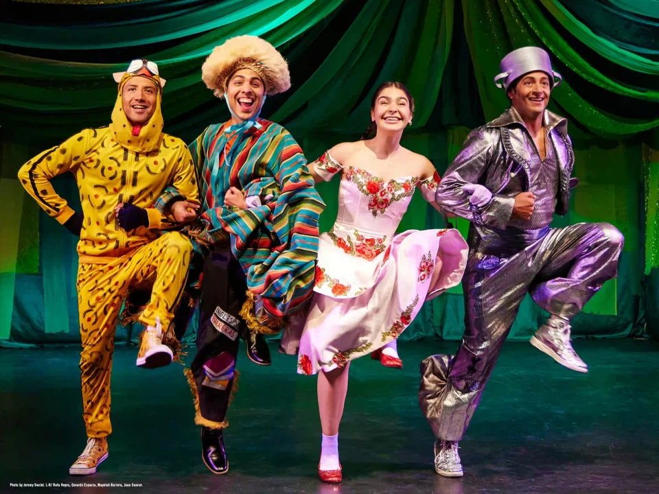 Four performers in colorful costumes, including a yellow lion, striped scarecrow, floral dress, and silver tin man, smile and kick their legs in unison on a stage with a green fabric backdrop.