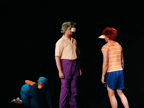 Three individuals are dressed in colorful costumes performing on stage; two stand facing each other, while the third crouches on the floor. The stage background is black.