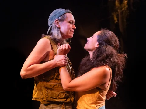 Two women stand close together on stage, looking into each other's eyes. One grips the other's shirt. The setting is dimly lit.