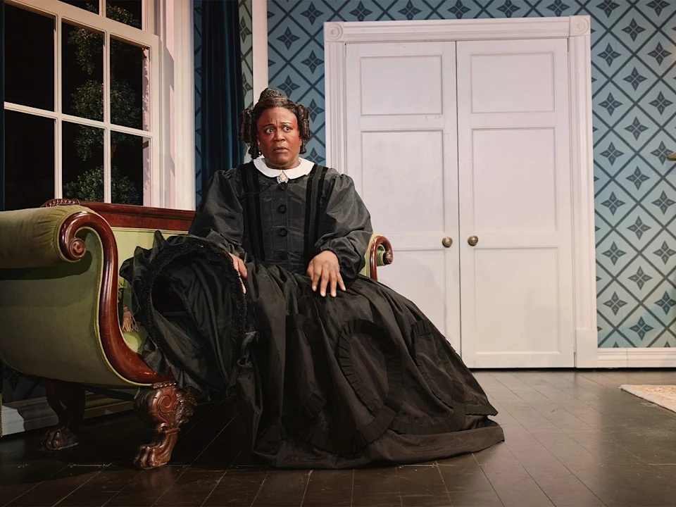 A person dressed in a formal black Victorian-style dress sits on a green couch in a room with patterned wallpaper and large white double doors.