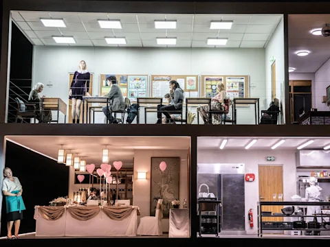 Production photo of Innocence in New York, showing a two-story set shows people in a classroom on the upper level and a decorated dining area and kitchen with staff on the lower level.