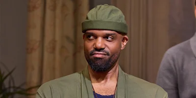 A person wearing a green hat and green jacket looks to the side with a skeptical expression indoors.