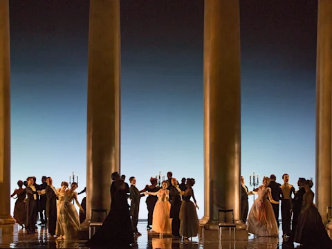 A group of people in formal attire dance in pairs between large columns, with candelabras in the background and a blue gradient backdrop.