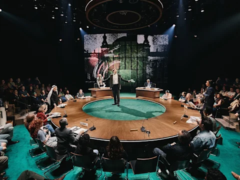 Production photo of Kyoto in New York, showing a person stands speaking in the center of a round stage surrounded by seated audience members, with three panelists at desks in the background.