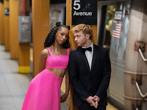 A woman in a bright pink outfit and a man in a black tuxedo stand next to a subway pillar marked "5 Avenue," with a subway train in the background.