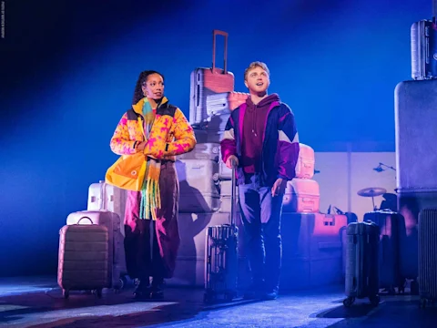 Two people stand on stage surrounded by stacked suitcases under blue lighting, both wearing colorful jackets and holding luggage.