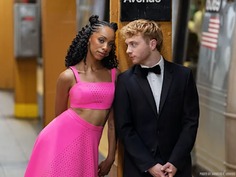 A woman in a bright pink crop top and skirt stands next to a man in a black tuxedo on a subway platform, with a train and an Avenue sign in the background.