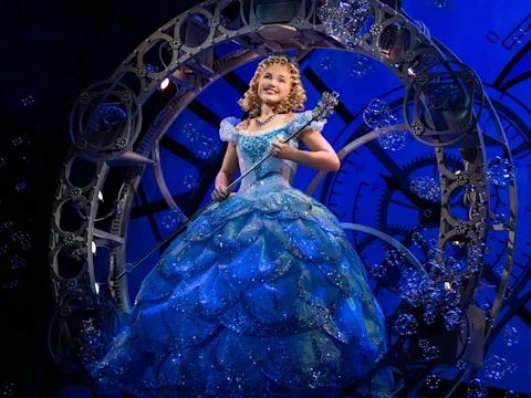 A performer in a blue, sparkling gown holds a wand and stands inside a circular set piece with gears, surrounded by stage bubbles and blue lighting.