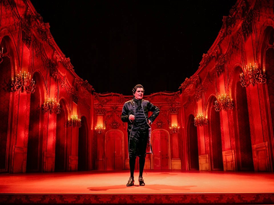 A man in period costume stands center stage in an ornate, red-lit theater set resembling a grand ballroom with chandeliers.