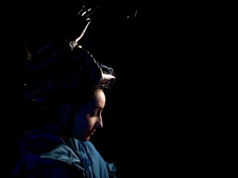 A person in profile wearing an elaborate, dark headpiece and blue clothing is partly illuminated against a black background.