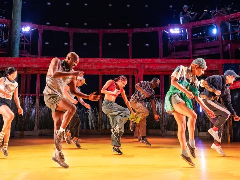 A group of dancers, dressed in casual, sporty attire, perform synchronized moves on a brightly lit stage with a rustic red background and illuminated balcony.