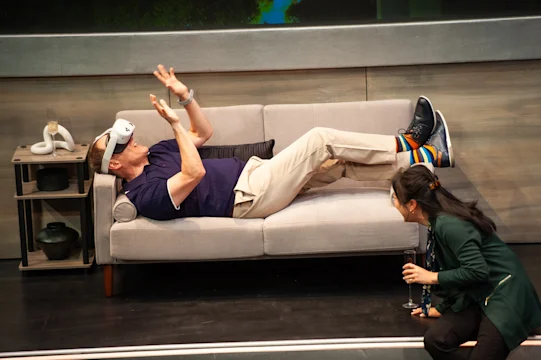 A man wearing a VR headset lies on a couch with his arms raised, while a woman kneels nearby holding a glass, looking at him.