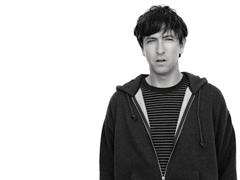 Nicholas Braun wearing a striped shirt and zip-up hoodie stands against a plain white background, looking toward the camera.