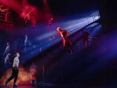 A person in a long coat is suspended in midair under dramatic stage lighting, while others watch from metal staircases and platforms in a dark, smoky theater setting.