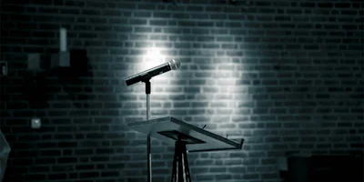 A microphone on a stand sits atop a podium in front of a brick wall, illuminated by focused lighting.