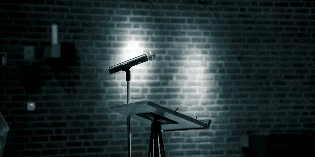 A microphone on a stand sits atop a podium in front of a brick wall, illuminated by focused lighting.