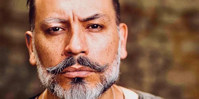Close-up of a middle-aged man with a styled mustache and beard, neutral expression, and blurred background.