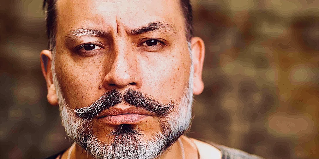 Close-up of a middle-aged man with a styled mustache and beard, neutral expression, and blurred background.