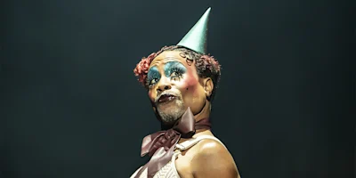 Person wearing a cone party hat, heavy theatrical makeup, and a brown ribbon bow around their neck, looking to the side with a dramatic expression against a dark background.