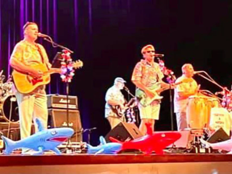 A band performs on stage with tropical decorations and inflatable sharks. The musicians wear tropical shirts and play various instruments, including guitars, congas, and drums.