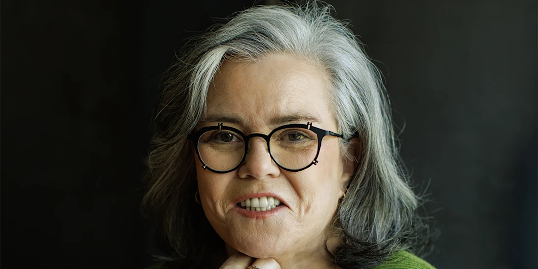 A woman with gray hair and black-rimmed glasses smiles slightly, resting her chin on her hand, against a dark background.