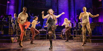 Performers energetically dance and sing on stage under purple lighting, with musicians and stage props in the background.