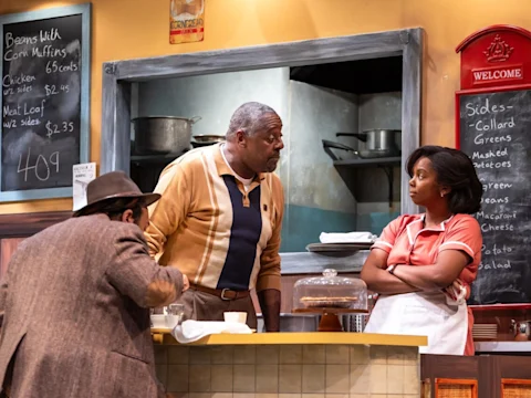 A man and woman have a conversation at a diner counter. Another man leans over eating. The menu board displays prices. Pots and a cake are visible in the background.