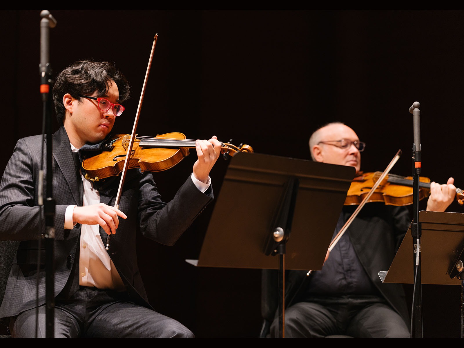 Two violinists in formal attire perform on stage, each reading sheet music from a stand, with microphones positioned nearby.