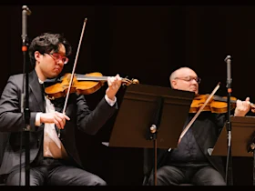 Two violinists in formal attire perform on stage, each reading sheet music from a stand, with microphones positioned nearby.