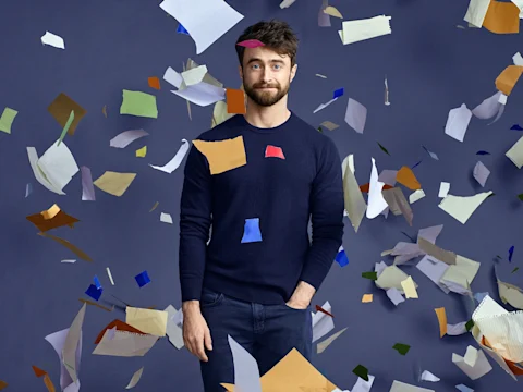 A man in a navy sweater stands against a gray backdrop as colorful paper sheets float around him in midair.