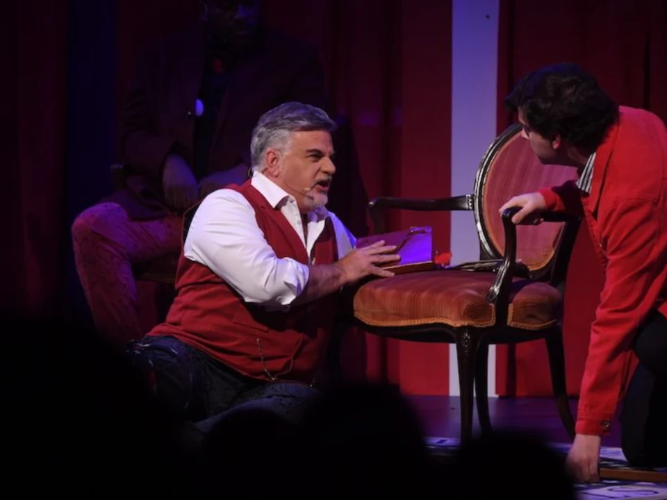 Two men in a performance scene. One man with gray hair is kneeling and holding a book, while another man in red is crouched next to a chair. A third person is partially visible in the background.